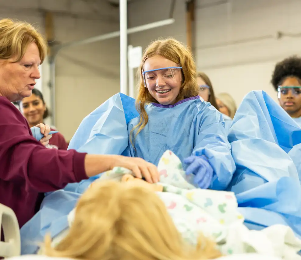 Student wearing safety glasses, scrubs, and gloves helps in mock labor and delivery simulation at MakerFest